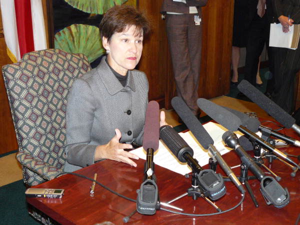 Alex Sink sitting at a wood desk surrounded by microphones